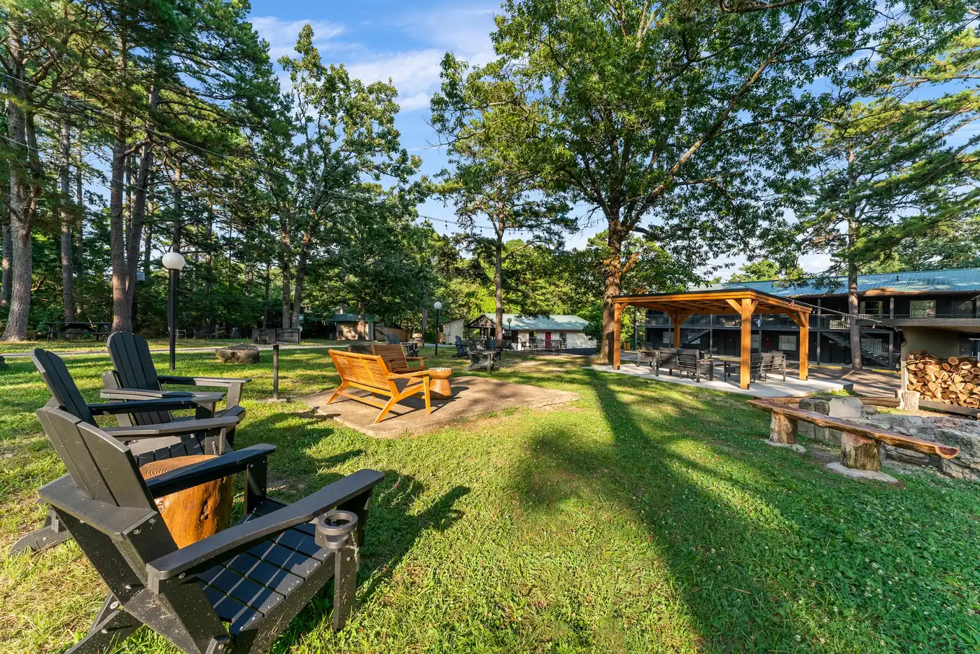 Lawn at Forest Springs Resort in Eureka Springs with Adirondack chairs, a stone fire ring with log benches and the covered outdoor lounge under tall pines