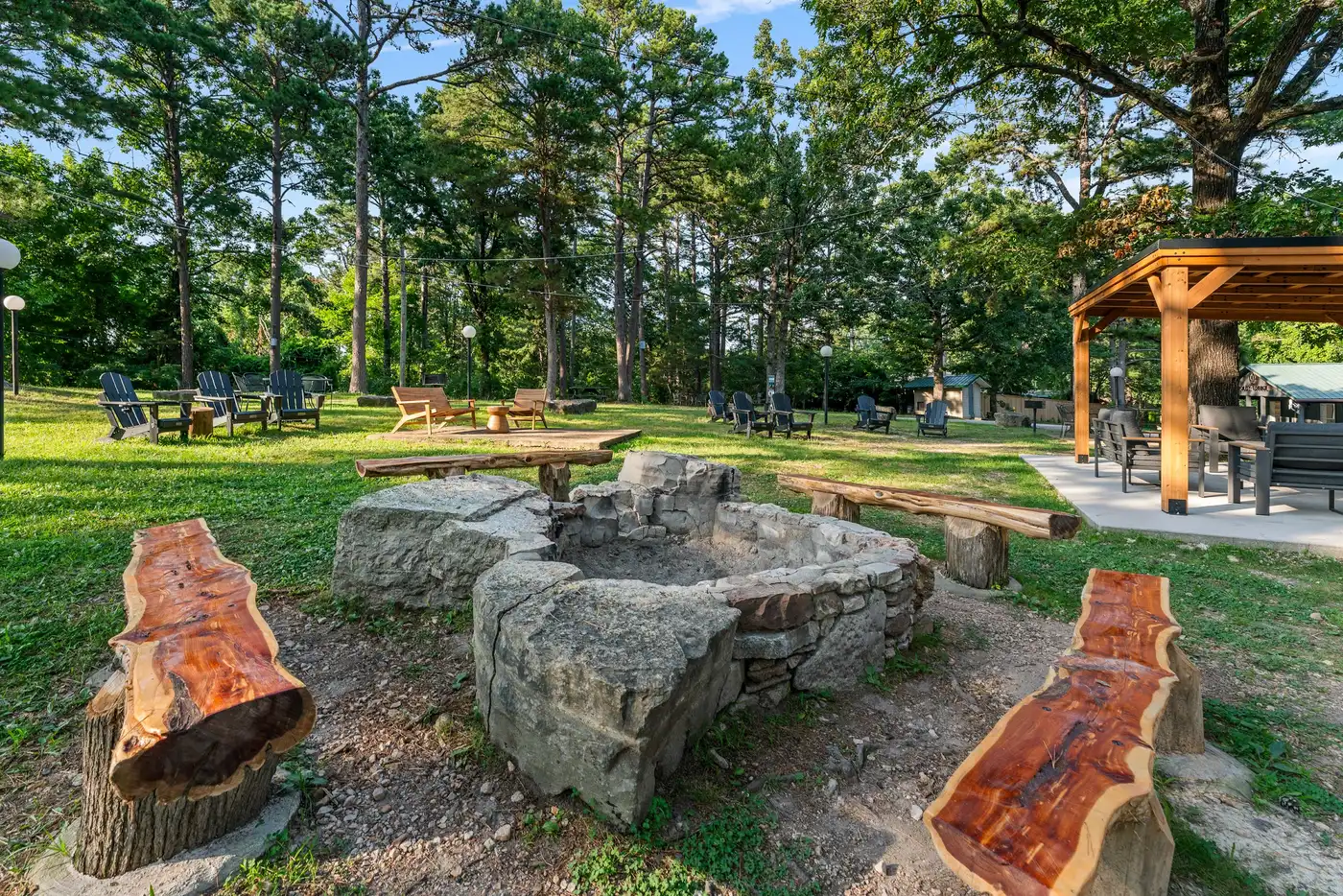 Stone fire ring ringed by cedar log benches on the lawn at Forest Springs Resort, with Adirondack chairs and the covered outdoor lounge behind